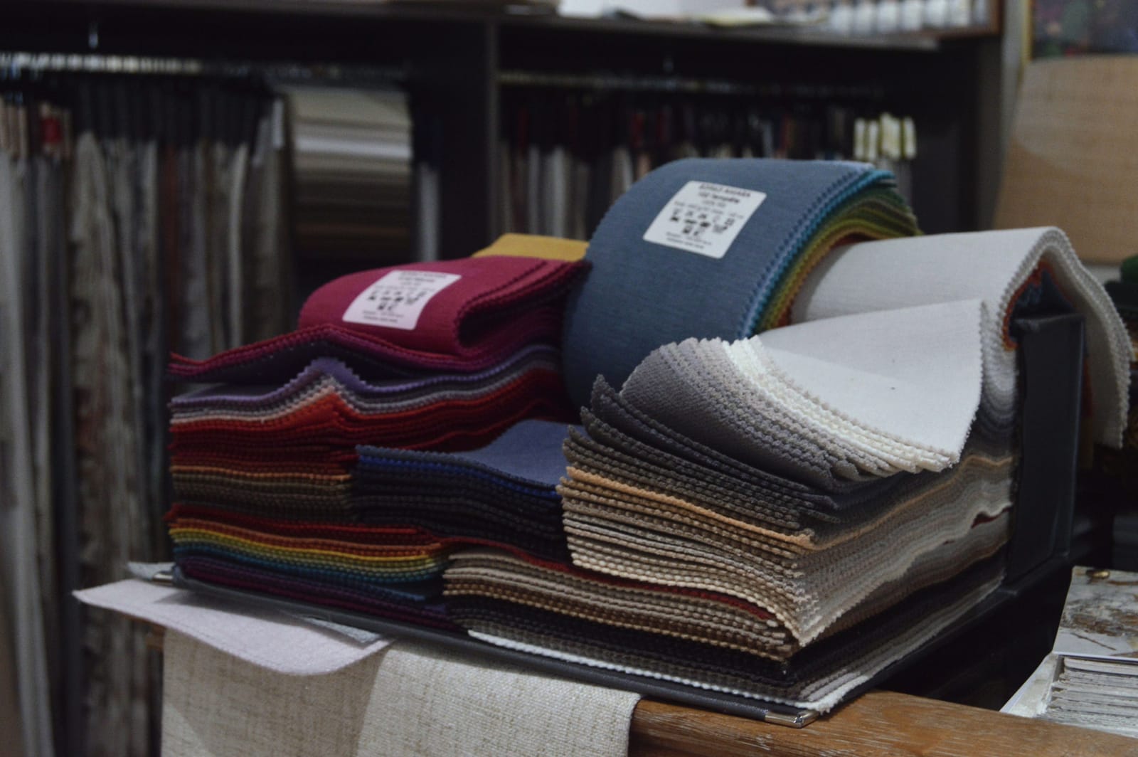Mostruários de tecidos em muitas cores numa mesa do showroom; ao fundo, mais catálogos pendurados
